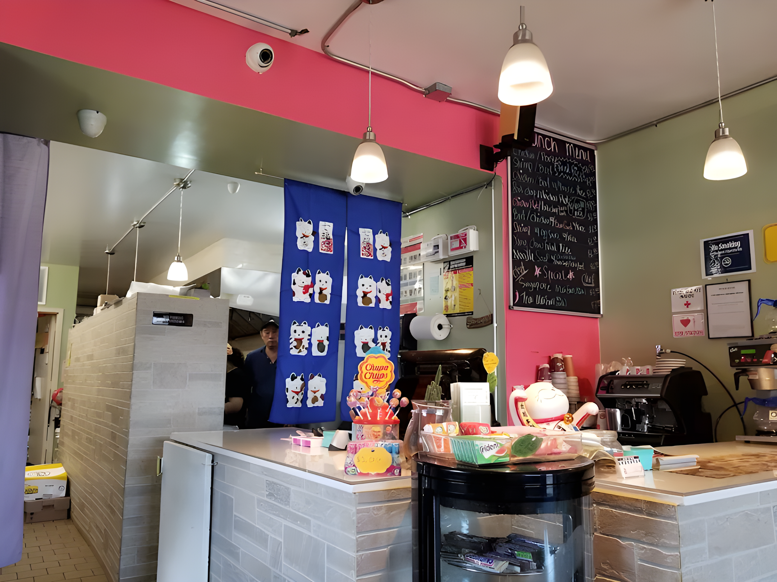 Cozy dessert shop interior with blue lucky cat banners, pink walls, and a counter displaying handmade Snowflake Crisps treats.